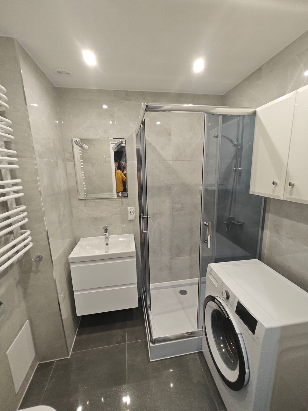 Nowoczesna łazienka z kabiną prysznicową, umywalką z szafką, grzejnikiem drabinkowym i pralką, wykończona szarymi płytkami w odcieniu marmuru, z odbiciem osoby w lustrze.