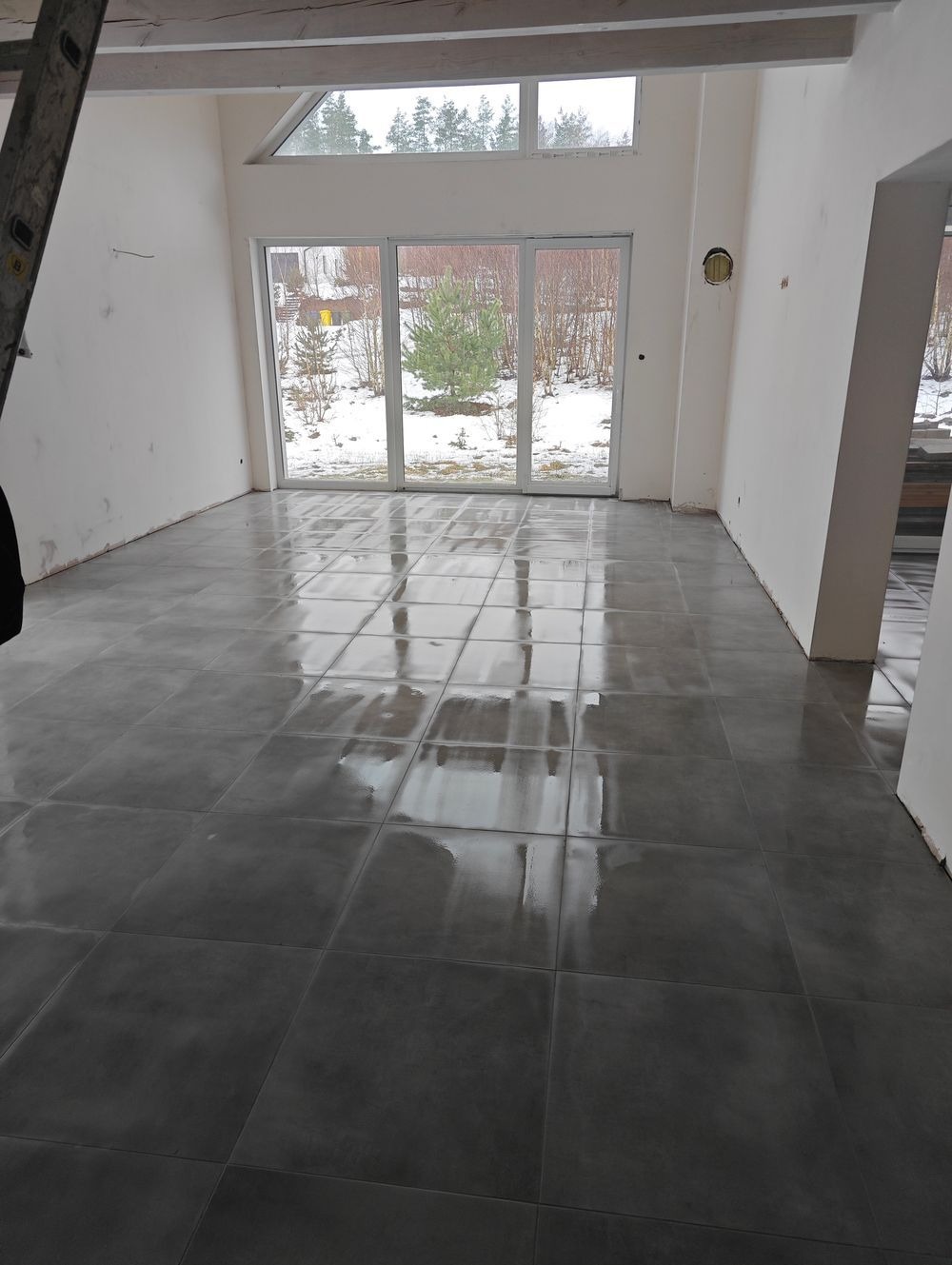 Świeżo ułożona, błyszcząca, ciemnoszara podłoga z płytek w przestronnym, jasnym pokoju z dużymi oknami tarasowymi, przez które widać zimowy krajobraz. Na ścianach widoczne ślady po pracach...