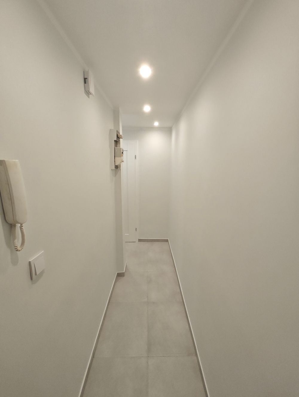 Wąski, jasny korytarz z szarymi płytkami podłogowymi, oświetlony punktowymi lampami sufitowymi, z białymi ścianami i widocznym domofonem po lewej stronie.