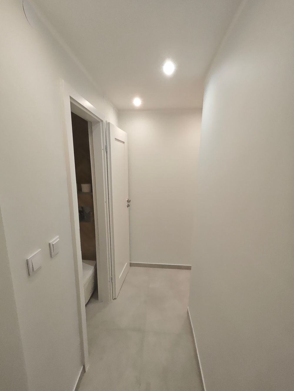 Korytarz z białymi ścianami, szarą podłogą i otwartymi drzwiami do łazienki z widoczną wanną, oświetlony trzema punktowymi lampami sufitowymi.