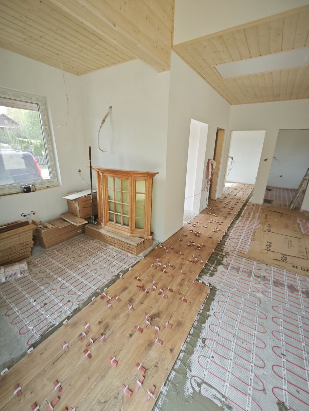 Montaż drewnopodobnych płytek na ogrzewaniu podłogowym w jasnym wnętrzu, widoczne płytki z dystansami, przewody grzewcze i deski sufitowe.