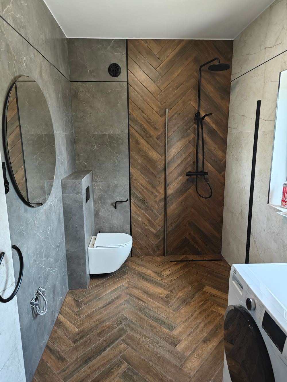 Nowoczesna łazienka z szarymi płytkami imitującymi marmur, drewnopodobną podłogą ułożoną w jodełkę, kabiną prysznicową walk-in z czarną armaturą i okrągłym lustrem.