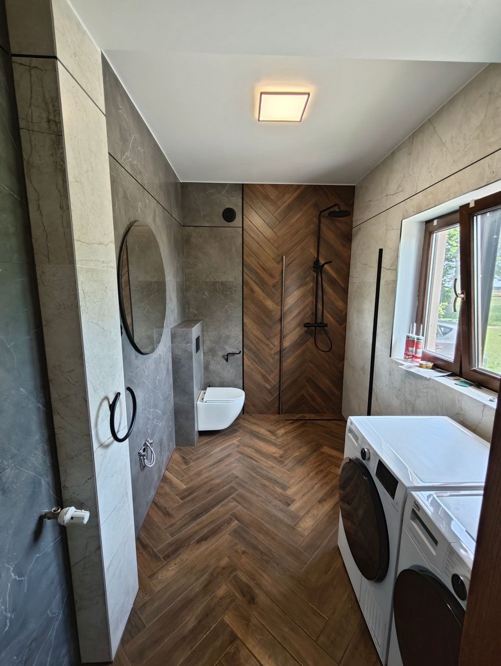 Nowoczesna łazienka z drewnianą podłogą ułożoną w jodełkę, szare płytki imitujące marmur na ścianach, okrągłe lustro, wisząca toaleta i pralka z suszarką.