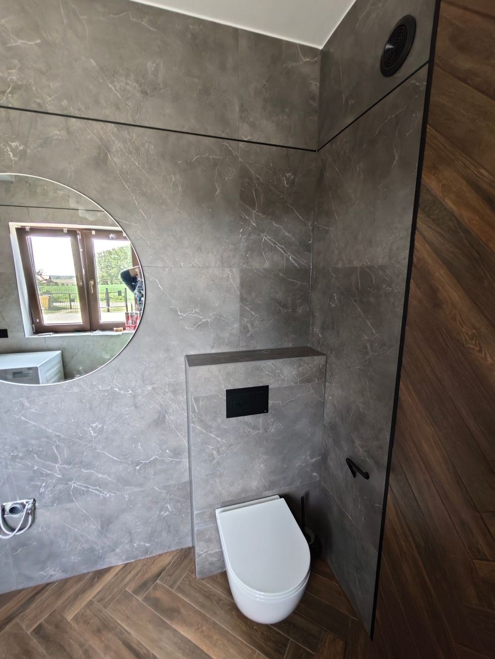 Nowoczesna łazienka z szarymi płytkami imitującymi marmur, podwieszana toaleta z czarnym przyciskiem, okrągłe lustro z widokiem na zewnątrz i parkiet ułożony we wzór jodełki.
