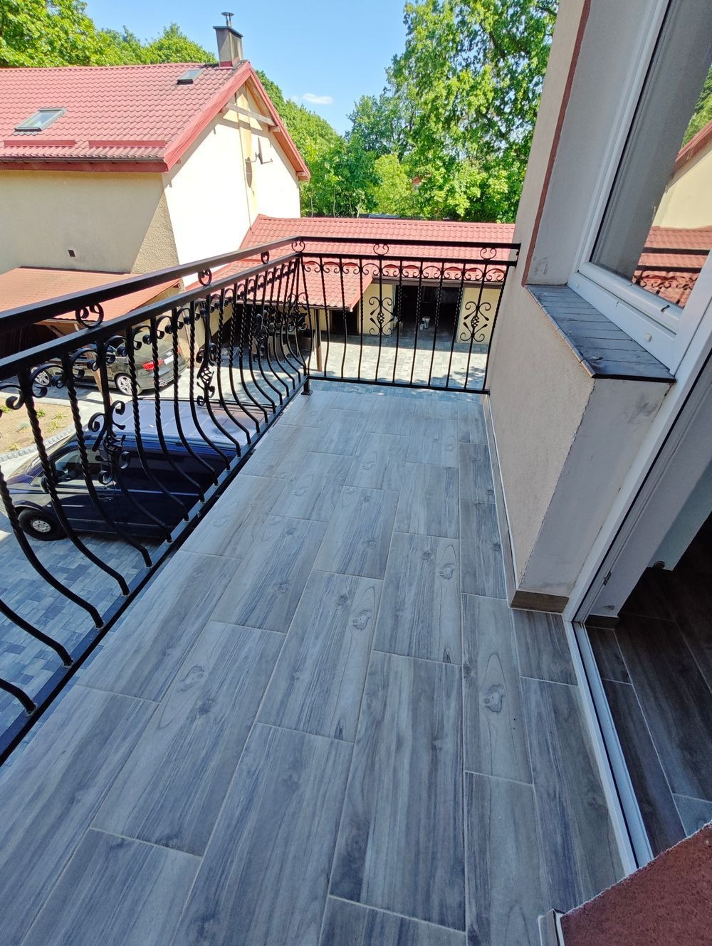 Wykończony balkon z szarymi płytkami imitującymi drewno, ozdobna metalowa balustrada, widok na budynek z czerwoną dachówką i zaparkowany samochód.