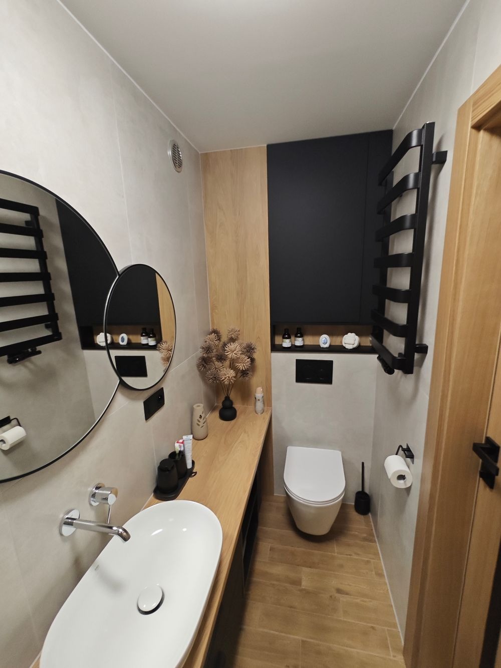 Nowoczesna łazienka z owalną umywalką nablatową, drewnianym blatem i podłogą w odcieniach drewna, czarnym grzejnikiem drabinkowym oraz okrągłym lustrem z podwójnym odbiciem.