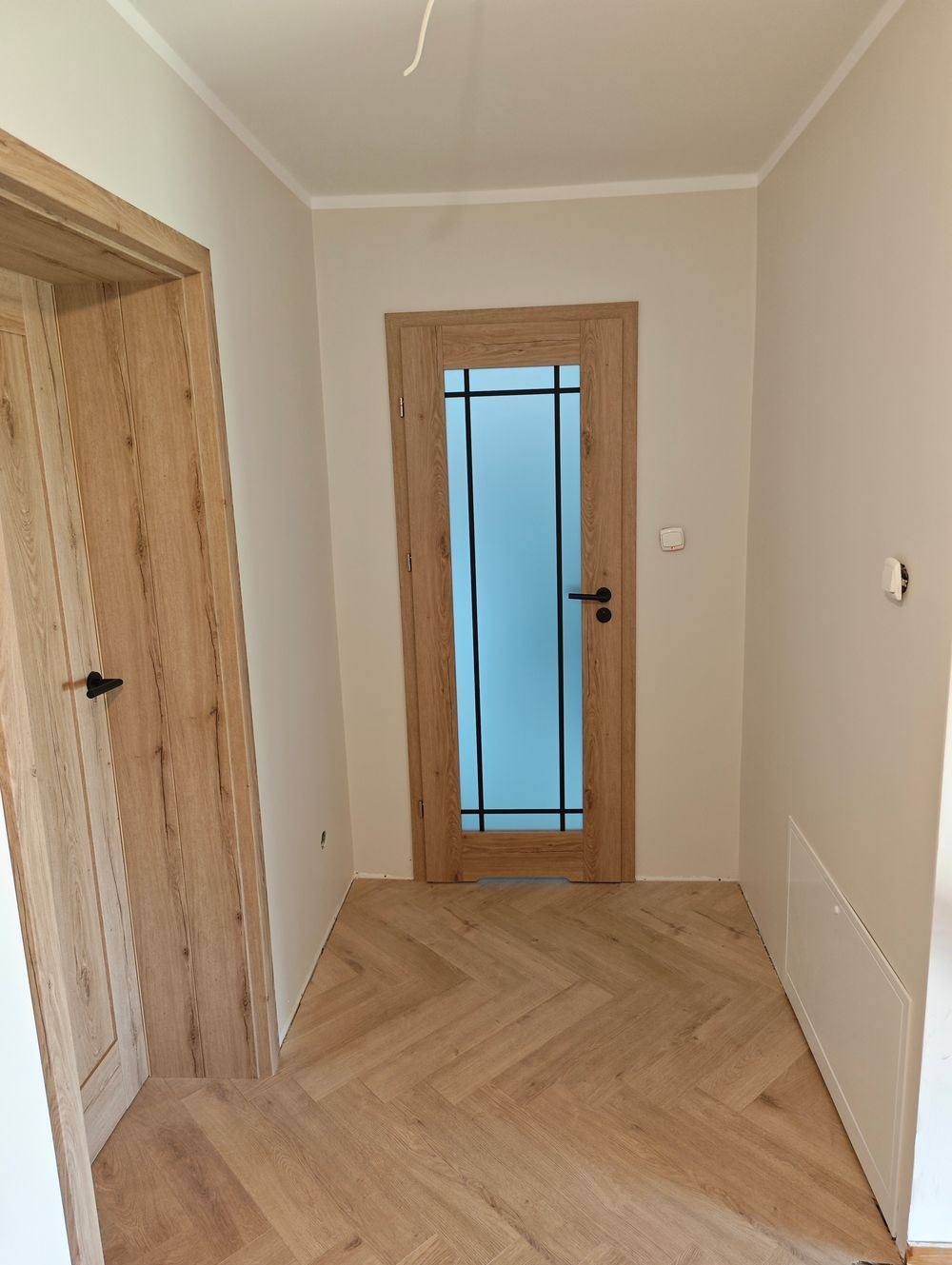 Korytarz z parkietem ułożonym w jodełkę, dwoje drewnianych drzwi, jedne z przeszkleniem, ściany w jasnym beżu.