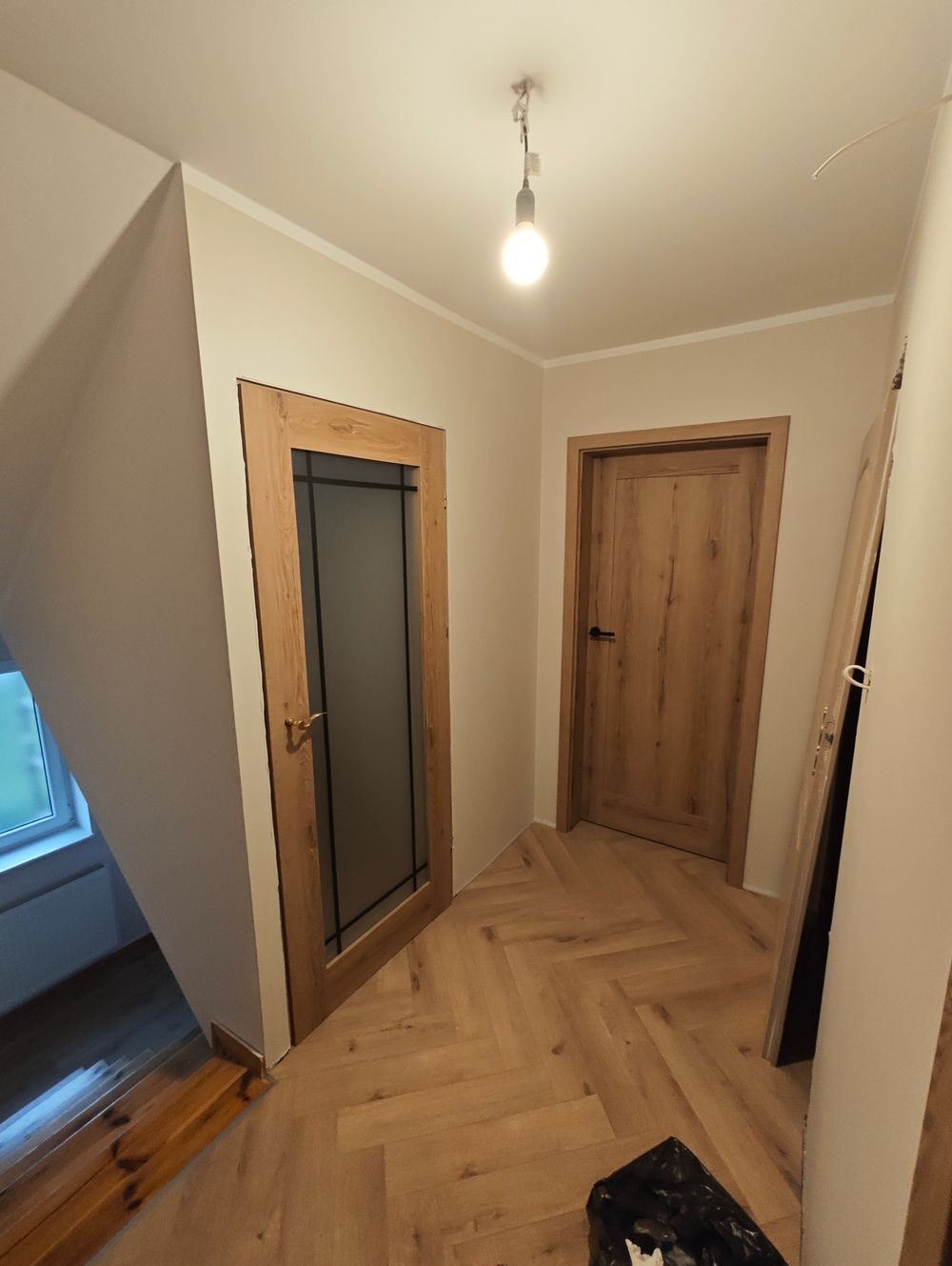 Korytarz po remoncie z ułożonym parkietem w jodełkę, widoczne dwie drewniane drzwi z ościeżnicami i fragment schodów przy oknie.