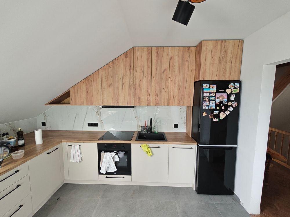 Kuchnia w skosach na poddaszu: zabudowa z drewnopodobnych szafek wiszących, białe szafki dolne z czarnymi uchwytami, marmurowy backsplash, czarna lodówka z magnesami, szare płytki podłogowe.