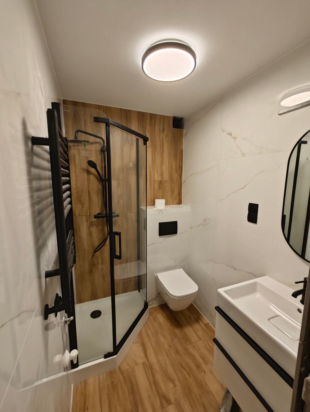 Nowoczesna łazienka z kabiną prysznicową typu walk-in z czarnymi profilami, umywalką nablatową, podwieszaną toaletą oraz płytkami imitującymi marmur i drewno.