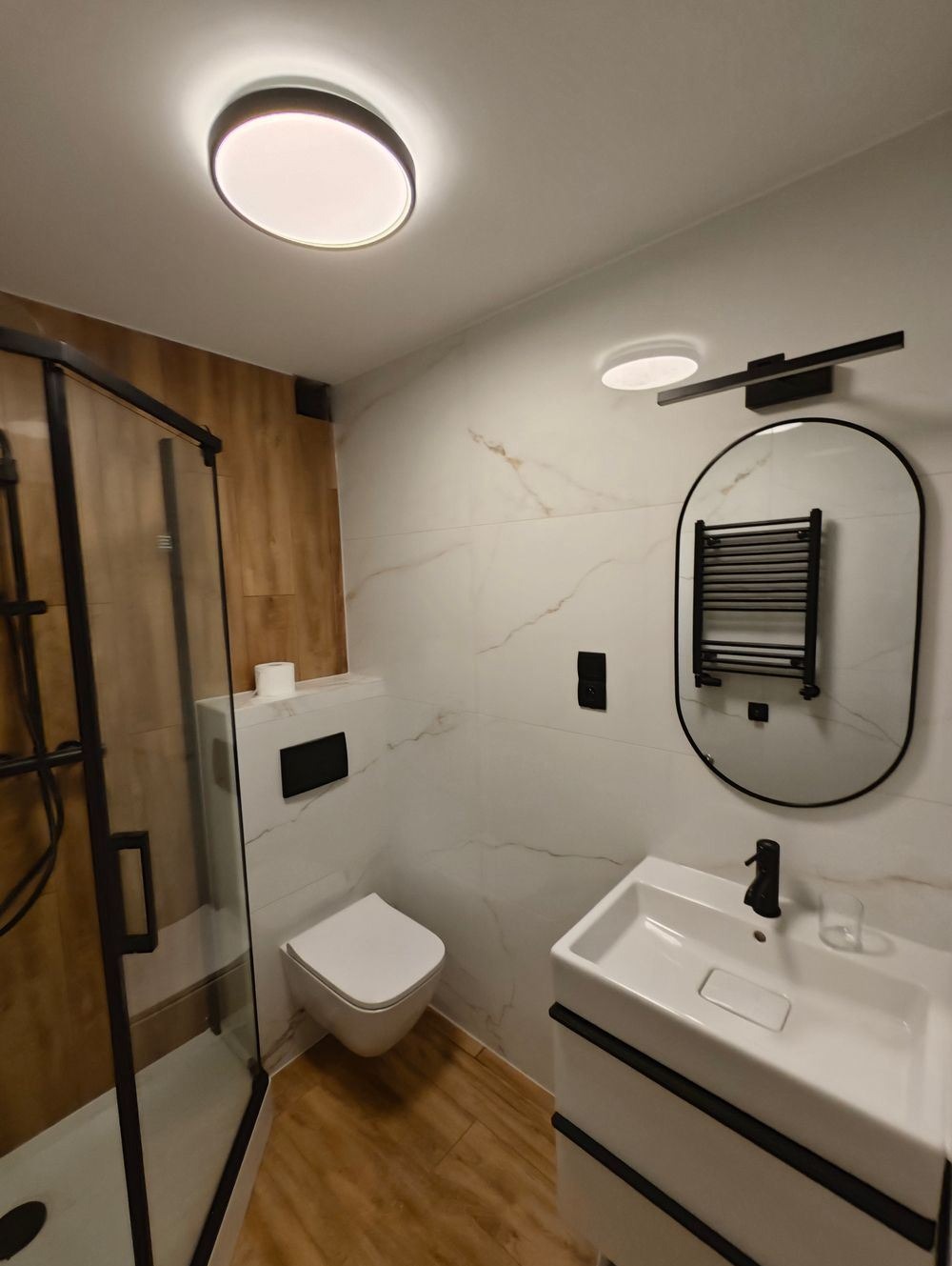 Nowoczesna łazienka z kabiną prysznicową z czarnymi profilami, podwieszaną toaletą, umywalką z szafką w stylu minimalistycznym, lustrem z grzejnikiem drabinkowym i oświetleniem LED.