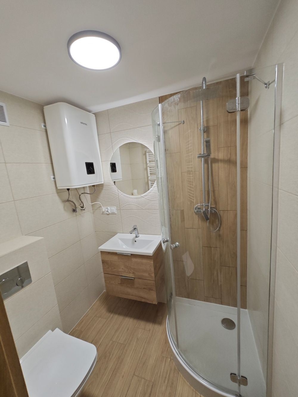 Jasna łazienka po renowacji z kabiną prysznicową, umywalką z drewnianą szafką, okrągłym lustrem i białym bojlerem Atlantic na ścianie.