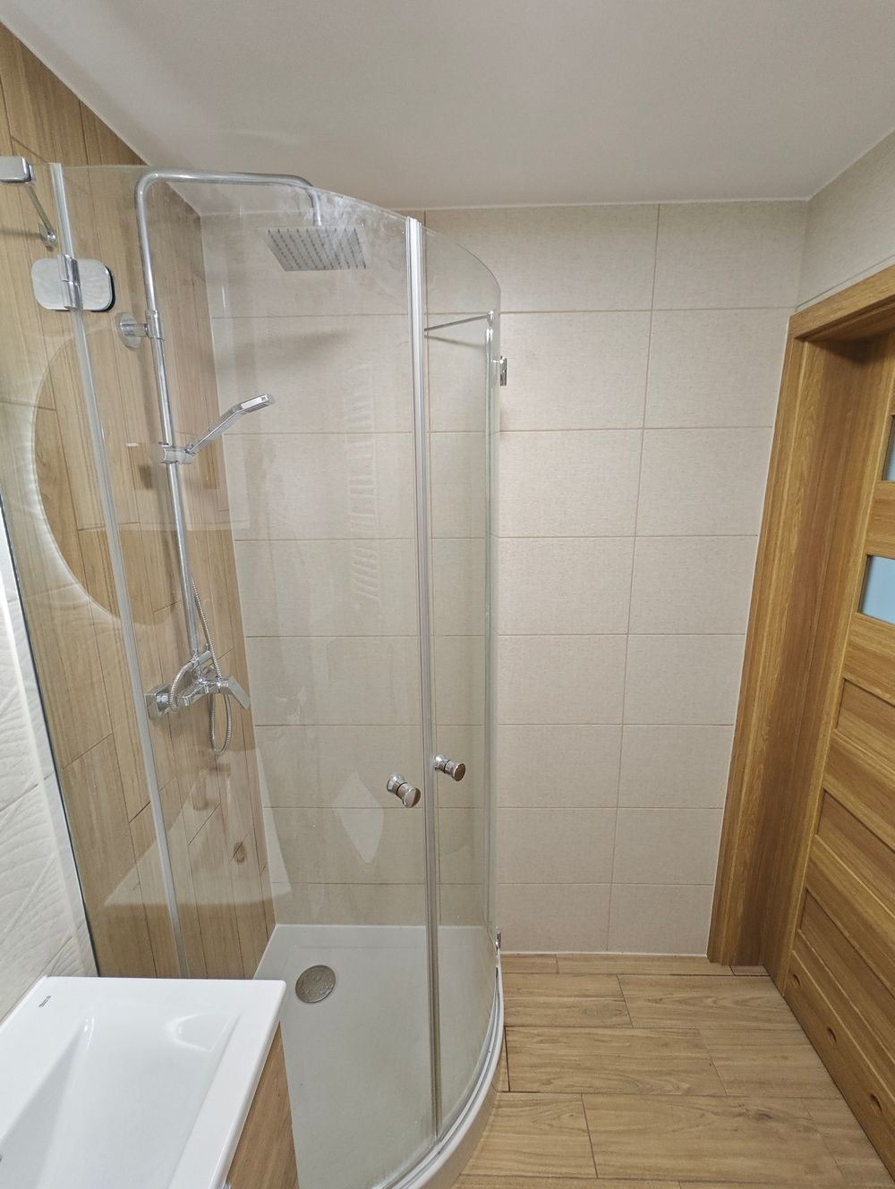 Nowoczesna kabina prysznicowa z chromowaną armaturą, umywalką z szafką w odcieniach drewna, jasne płytki ścienne i podłogowe imitujące drewno, widok z boku na wnętrze łazienki