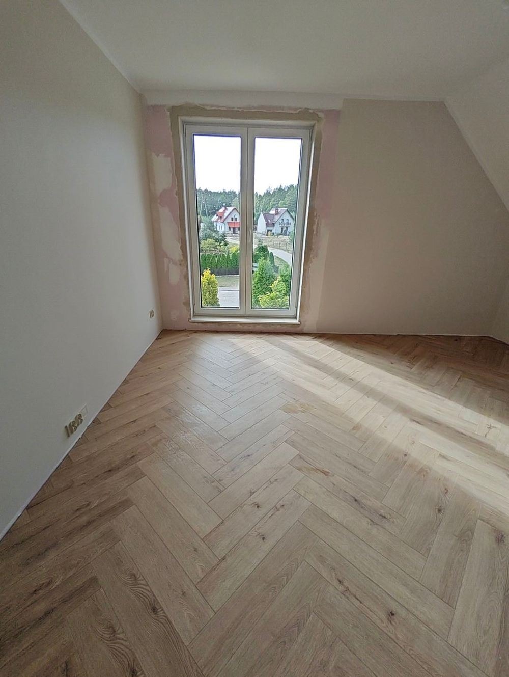 Jasny parkiet ułożony we wzór jodełki w pokoju z oknem, ściany przygotowane do malowania, widok na domy za oknem.