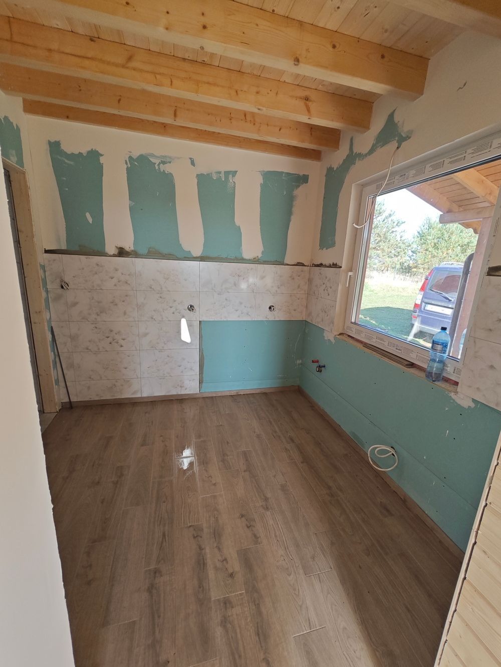 Wnętrze łazienki w trakcie remontu: ułożone płytki na ścianie, panele podłogowe w odcieniach brązu, płyty karton-gips na górze ściany i nad oknem, drewniany sufit z belkami, widoczna butelka wody...