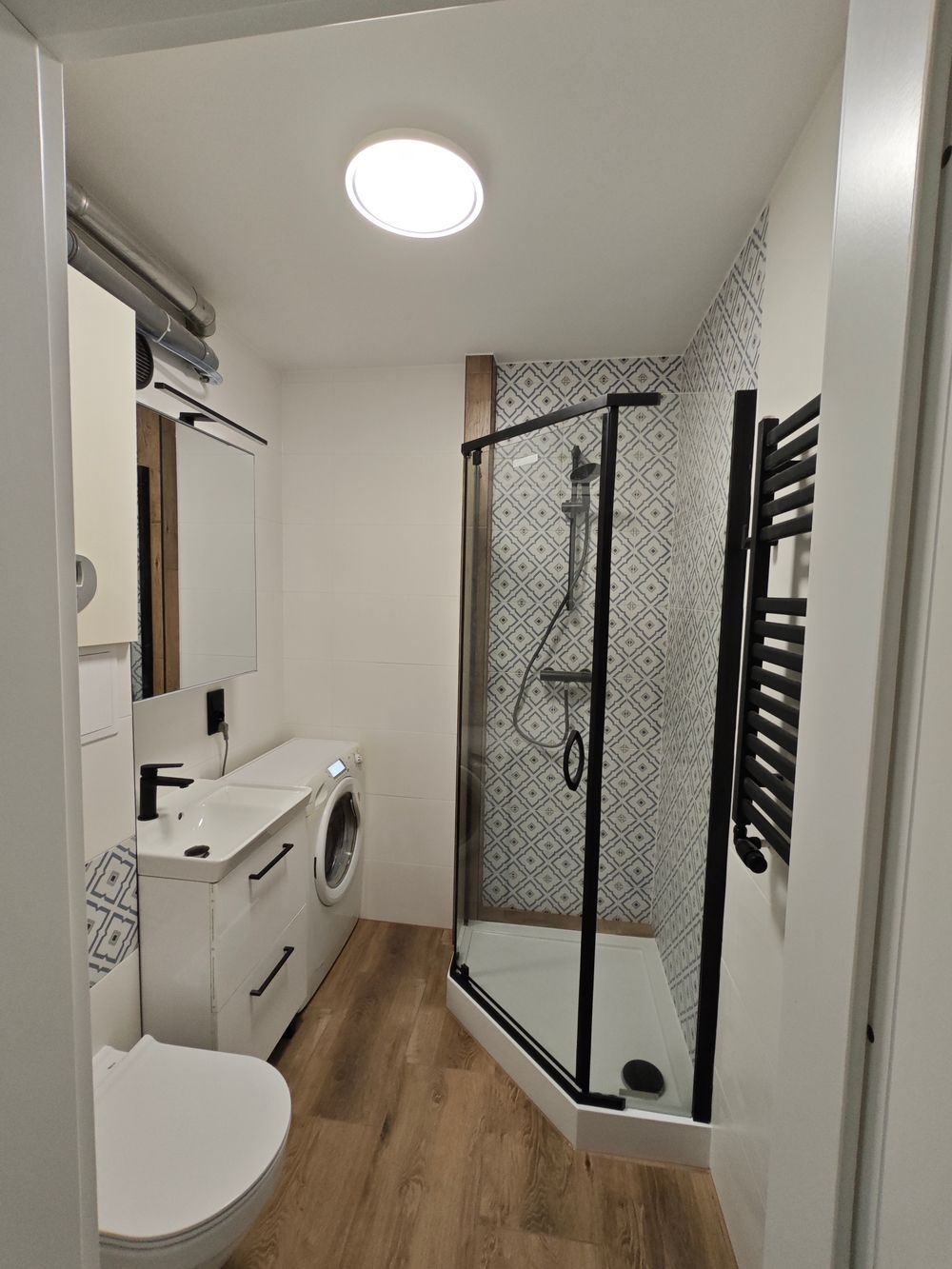 Nowoczesna łazienka z kabiną prysznicową typu walk-in z czarnymi profilami, umywalką z szafką, pralką i grzejnikiem drabinkowym w kolorze czarnym, wykończona płytkami w geometryczne wzory.