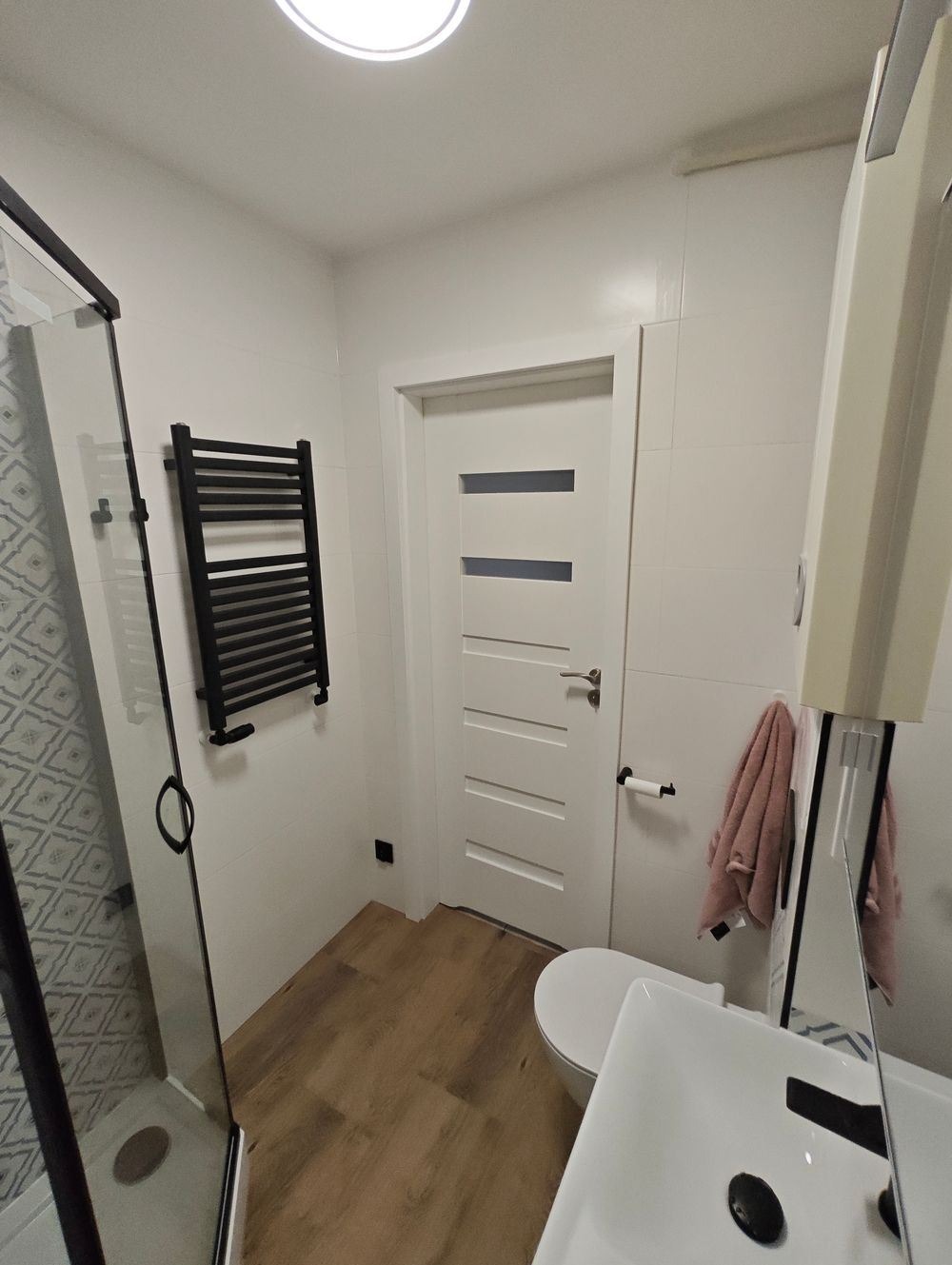 Nowoczesna łazienka z kabiną prysznicową z czarnymi profilami, drewnopodobną podłogą, białymi płytkami i grzejnikiem drabinkowym w kolorze czarnym.