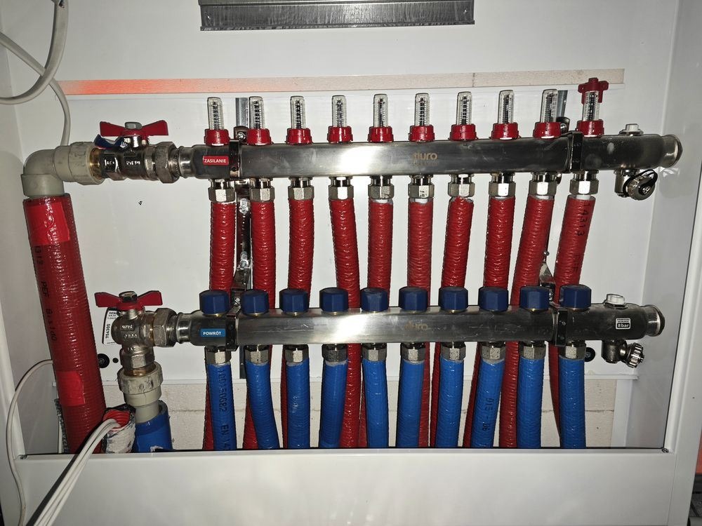 Rozdzielacz instalacji centralnego ogrzewania z czerwonymi i niebieskimi rurami w białej szafce, widoczne zawory i liczniki przepływu.