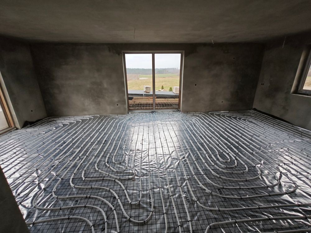 Szare wnętrze w trakcie montażu ogrzewania podłogowego z widocznymi rurami na czarnej siatce, surowe ściany w tle i okno z widokiem na zewnątrz.