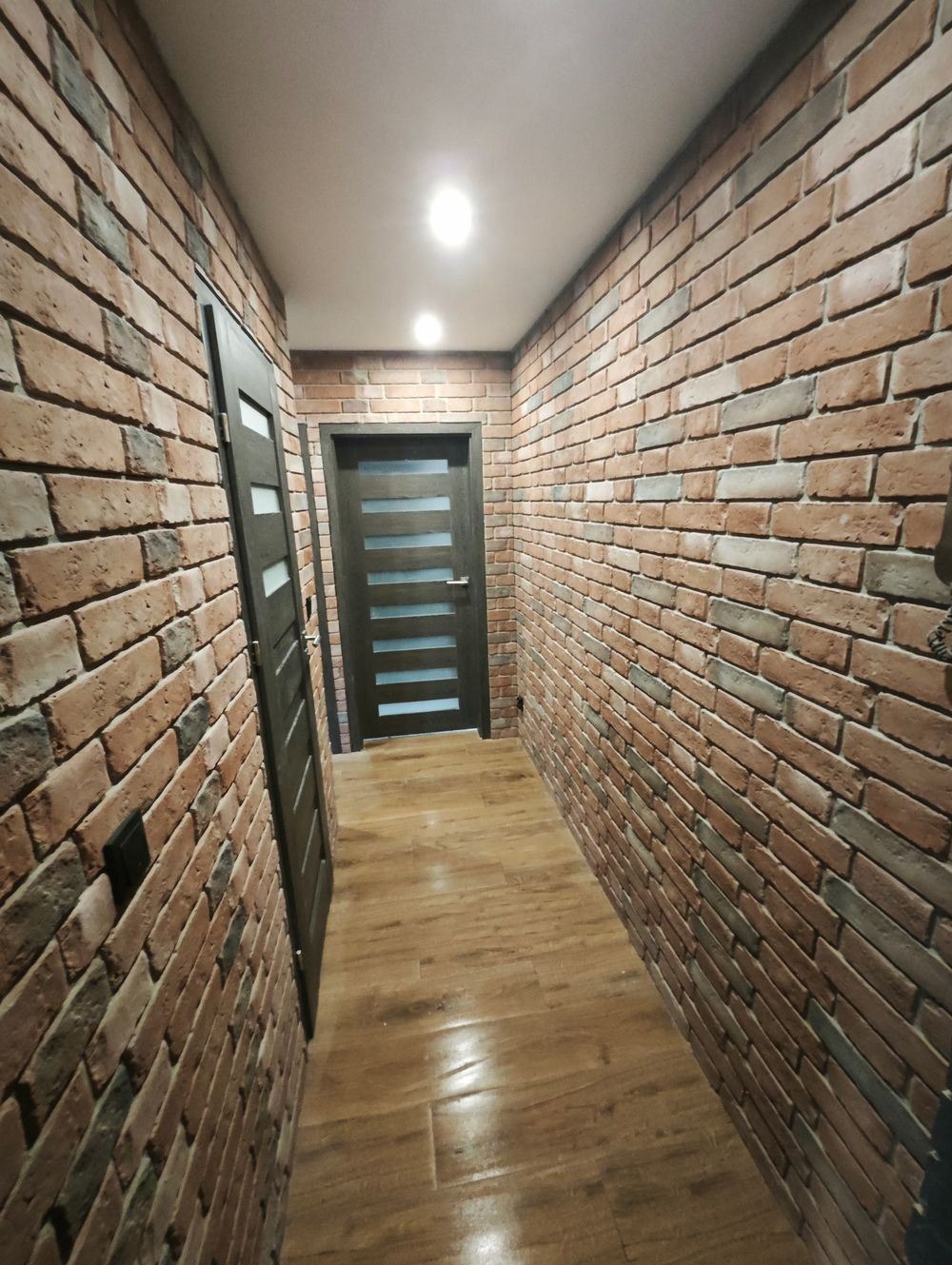 Wąski korytarz z wykończeniem ścian imitującym czerwoną cegłę, podłoga wyłożona panelami imitującymi drewno, widoczne dwie ciemne drewniane drzwi z przeszkleniami.