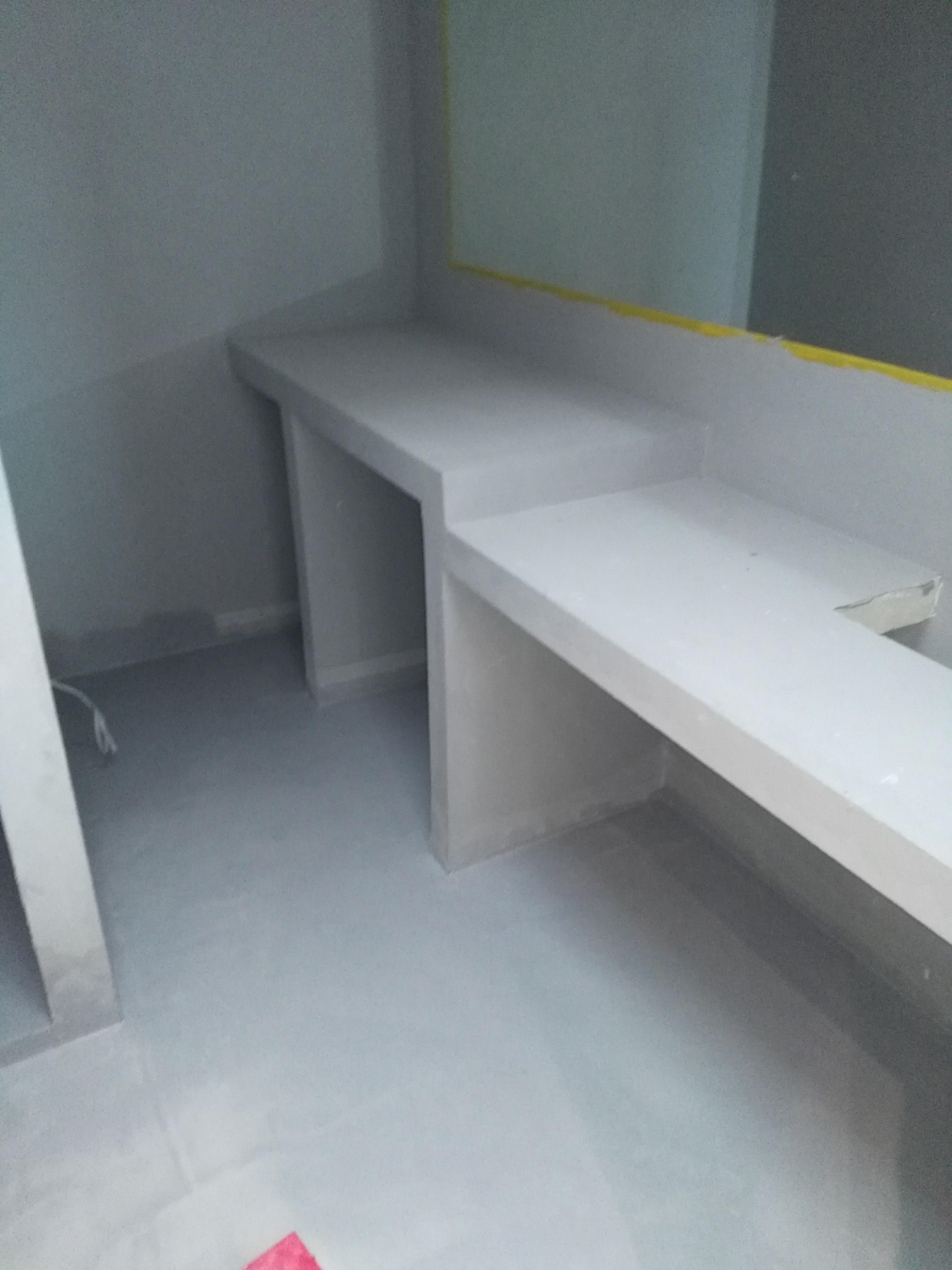 Surowy stan łazienki z przygotowaną pod zabudowę umywalki, widoczna szara ściana, podłoga i fragment lustra zabezpieczonego żółtą taśmą.