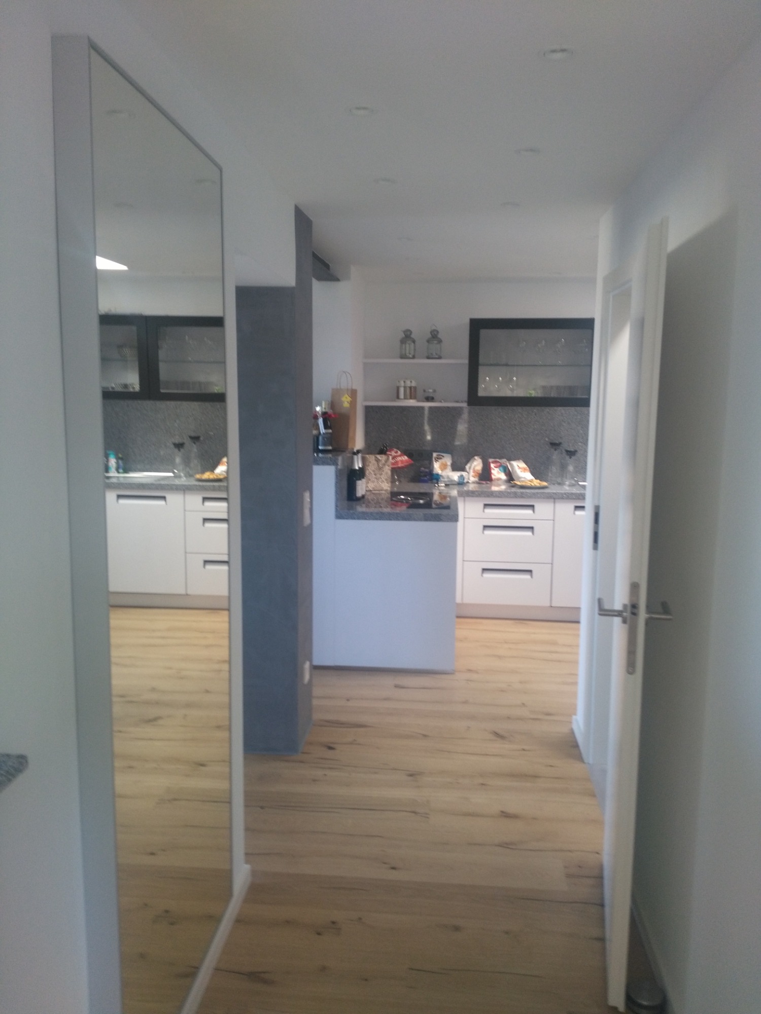 Wnętrze mieszkania z widoczną kuchnią w perspektywie korytarza, z lustrem po lewej stronie odbijającym część kuchni i drewnianą podłogą.