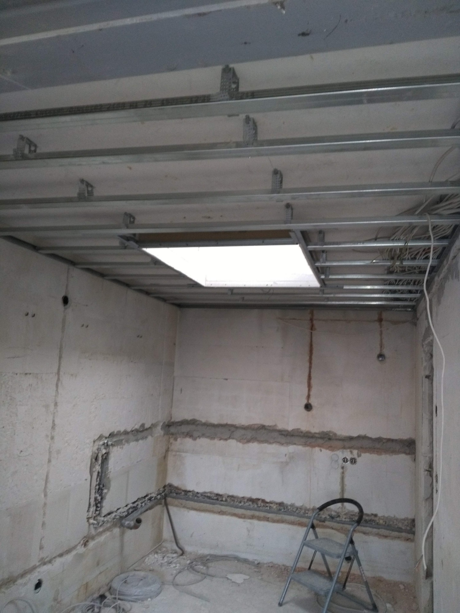 Wnętrze w trakcie prac: widoczna metalowa konstrukcja sufitu podwieszanego z oświetleniem, przygotowane ściany z instalacją elektryczną i drabina.