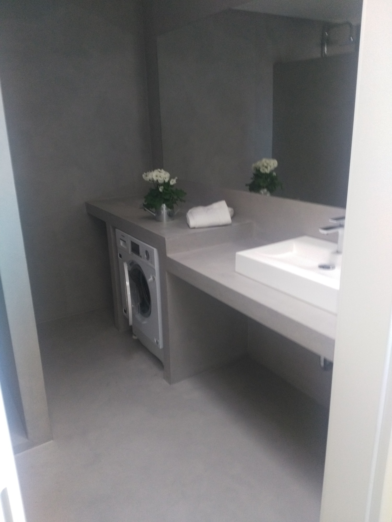 Minimalistyczna łazienka w odcieniach szarości z pralką w zabudowie pod blatem umywalki, biała umywalka i kwiaty w metalowych osłonkach.