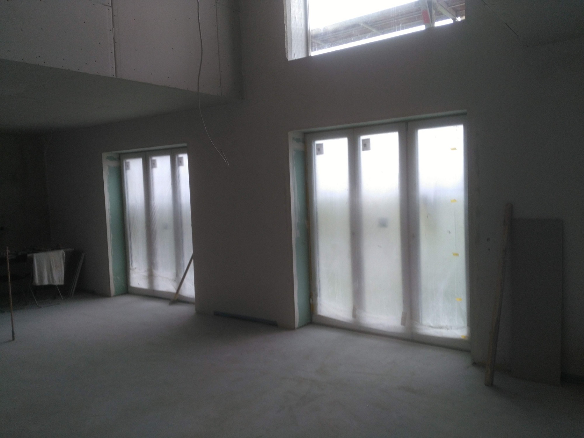 Surowe wnętrze z dwoma oknami zabezpieczonymi folią, widoczne niedokończone ściany z płyt gipsowo-kartonowych i surowa podłoga, przygotowane do dalszych prac wykończeniowych.