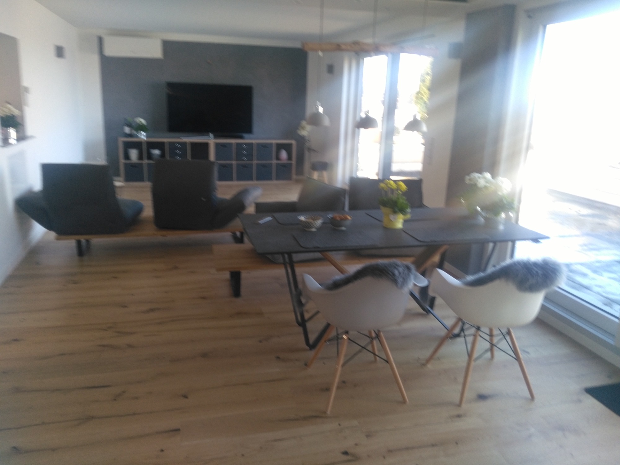 Jasna, drewniana podłoga w salonie z szarym stołem, krzesłami z futerkami, i minimalistycznym wystrojem wnętrza, w tle telewizor na ścianie.