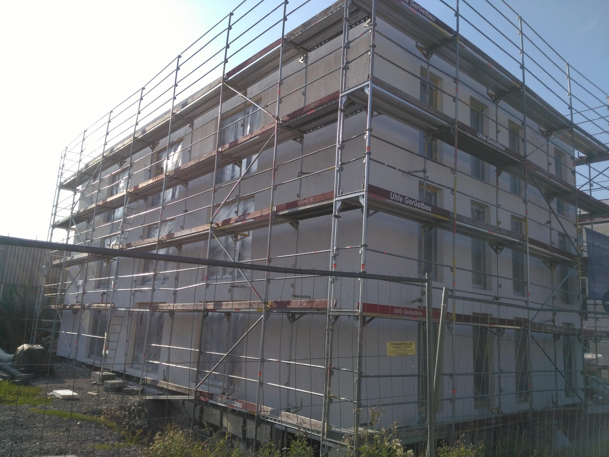 Budynek w trakcie prac elewacyjnych, osłonięty rusztowaniami z widocznymi oknami zabezpieczonymi folią. Na rusztowaniu widoczne oznaczenia producenta.