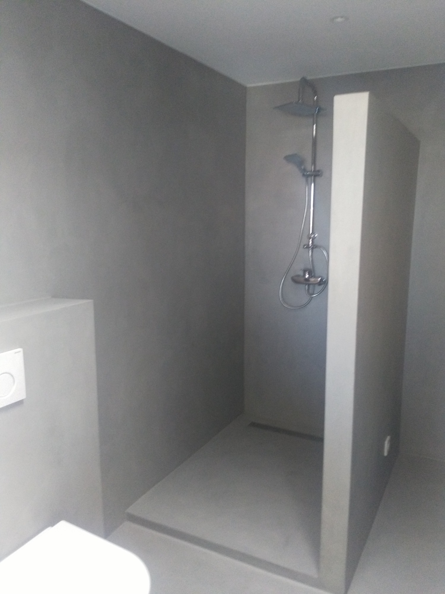 Minimalistyczna łazienka z kabiną prysznicową typu walk-in, wykończona w odcieniach szarości, z widoczną armaturą i przyciskiem spłuczki.