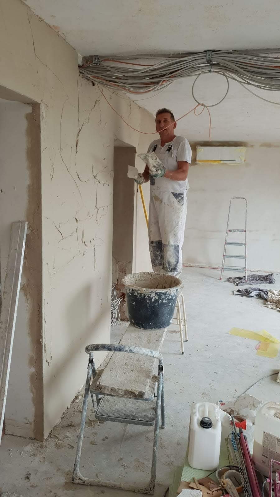 Mężczyzna w roboczym kombinezonie, stojąc na podeście, nakłada gładź na ścianę w pomieszczeniu z widocznymi przewodami elektrycznymi pod sufitem i drabinką w tle.