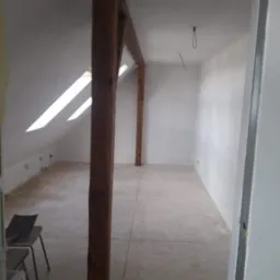 Poddasze w trakcie adaptacji: widoczne drewniane elementy konstrukcyjne dachu, skosy z oknami dachowymi, surowe ściany pomalowane na biało, częściowo położona podłoga z płyt, krzesło i narzędzia...