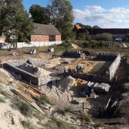 Fundamenty domu w trakcie budowy, wznoszone z szarej cegły, widoczny murarz w zielonym kasku i taczka, teren budowy z piaskiem i narzędziami, w tle zabudowa wiejska i błękitne niebo.