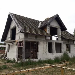 Niedokończony dom z częściowo pokrytym dachem, widoczne krokwie i dachówka, ściany z pustaków, niezadaszony balkon, rusztowania przy oknach, teren zarośnięty trawą.