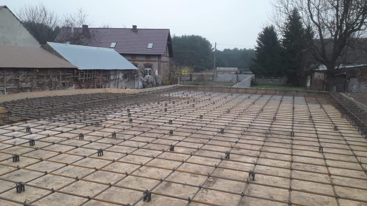 Zbrojenie płyty betonowej na deskowaniu, widoczne stalowe pręty ułożone w kratę z dystansami, w tle budynki gospodarcze i drzewa w pochmurny dzień.