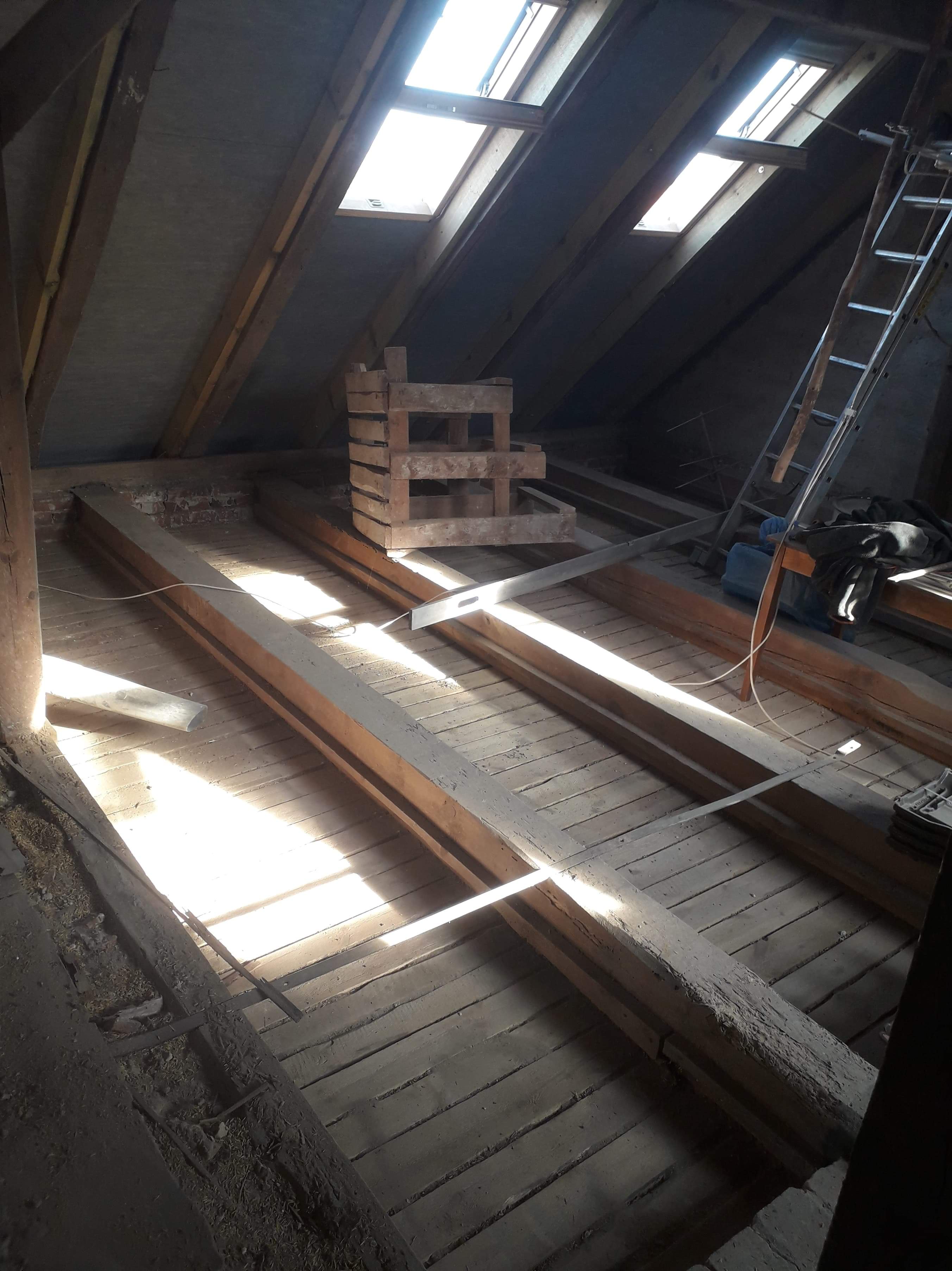 Wnętrze poddasza z drewnianą konstrukcją więźby dachowej w trakcie budowy, widoczne belki, deski podłogowe, drabina i okna dachowe wpadające światło.