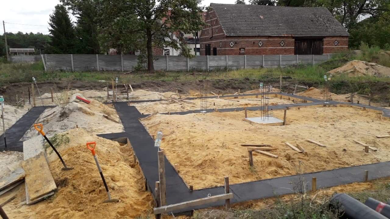 Przygotowanie fundamentów pod budowę domu jednorodzinnego: wykopane rowy zalane betonem, wystające pręty zbrojeniowe, w tle ceglany budynek gospodarczy i drewniany płot.