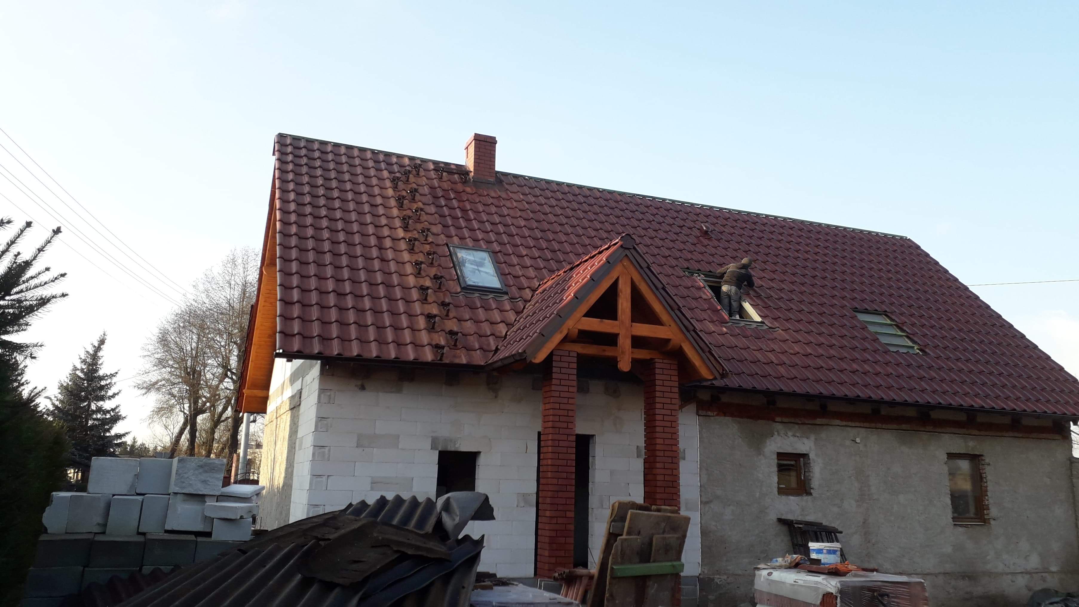 Dom w trakcie budowy z czerwoną dachówką, widoczny pracownik montujący okno dachowe i sterta bloczków betonowych przed budynkiem.