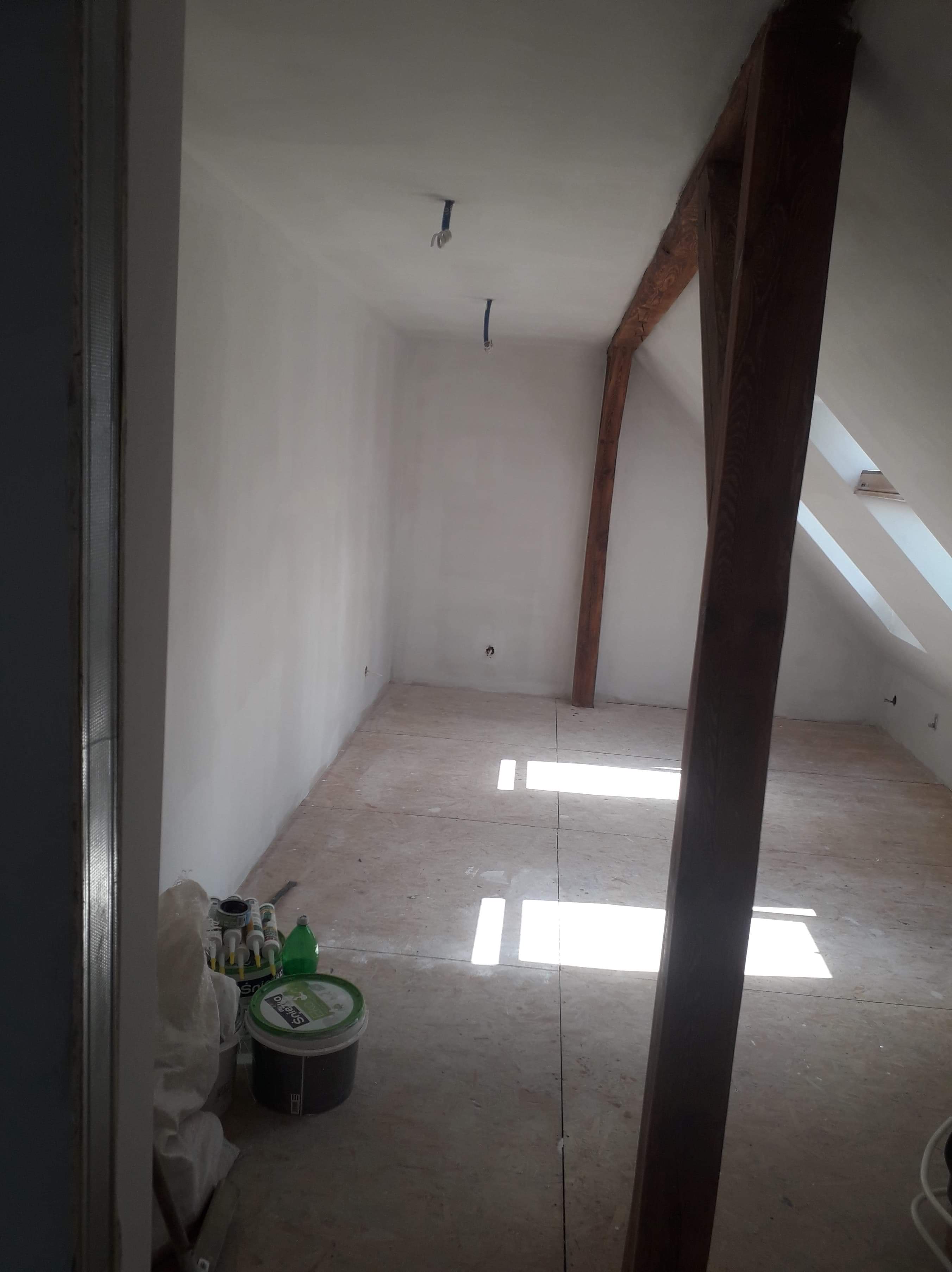 Poddasze w trakcie remontu z surowymi ścianami, widoczną więźbą dachową i materiałami budowlanymi w rogu pomieszczenia.