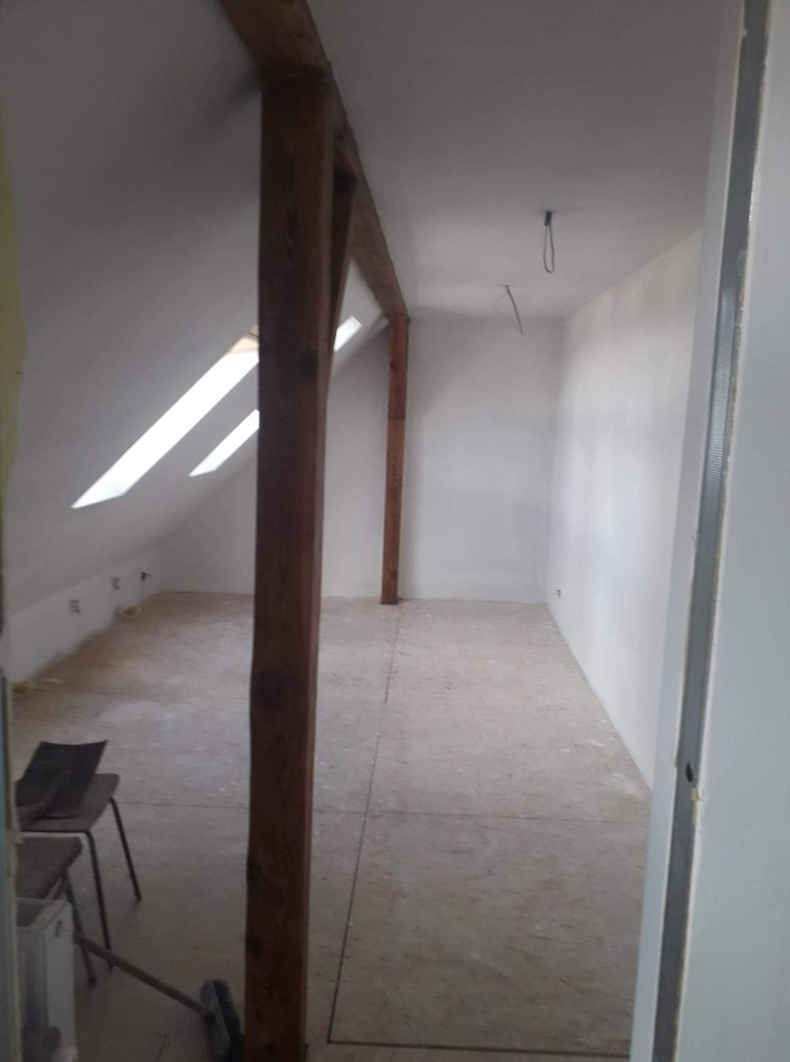 Poddasze w trakcie adaptacji: widoczne drewniane elementy konstrukcyjne dachu, skosy z oknami dachowymi, surowe ściany pomalowane na biało, częściowo położona podłoga z płyt, krzesło i narzędzia...