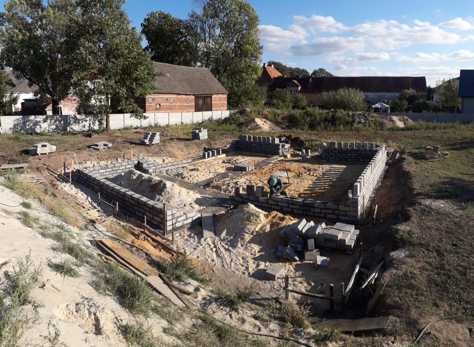 Fundamenty domu w trakcie budowy, wznoszone z szarej cegły, widoczny murarz w zielonym kasku i taczka, teren budowy z piaskiem i narzędziami, w tle zabudowa wiejska i błękitne niebo.