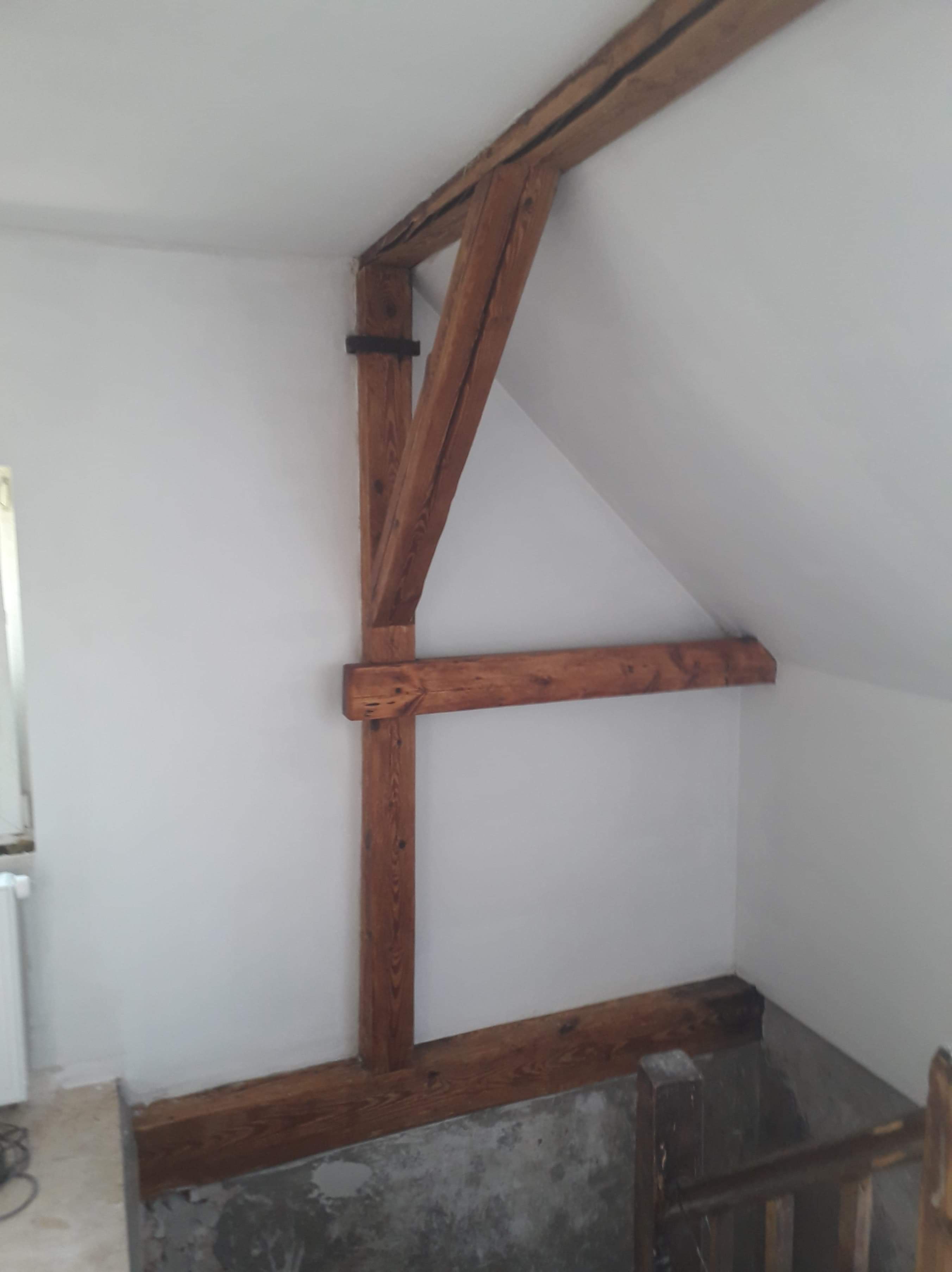 Wnętrze poddasza z elementami drewnianej konstrukcji nośnej dachu, widoczne słupy i belki w kolorze ciemnego brązu na tle białych ścian, fragmentarycznie wykończone podłoże.