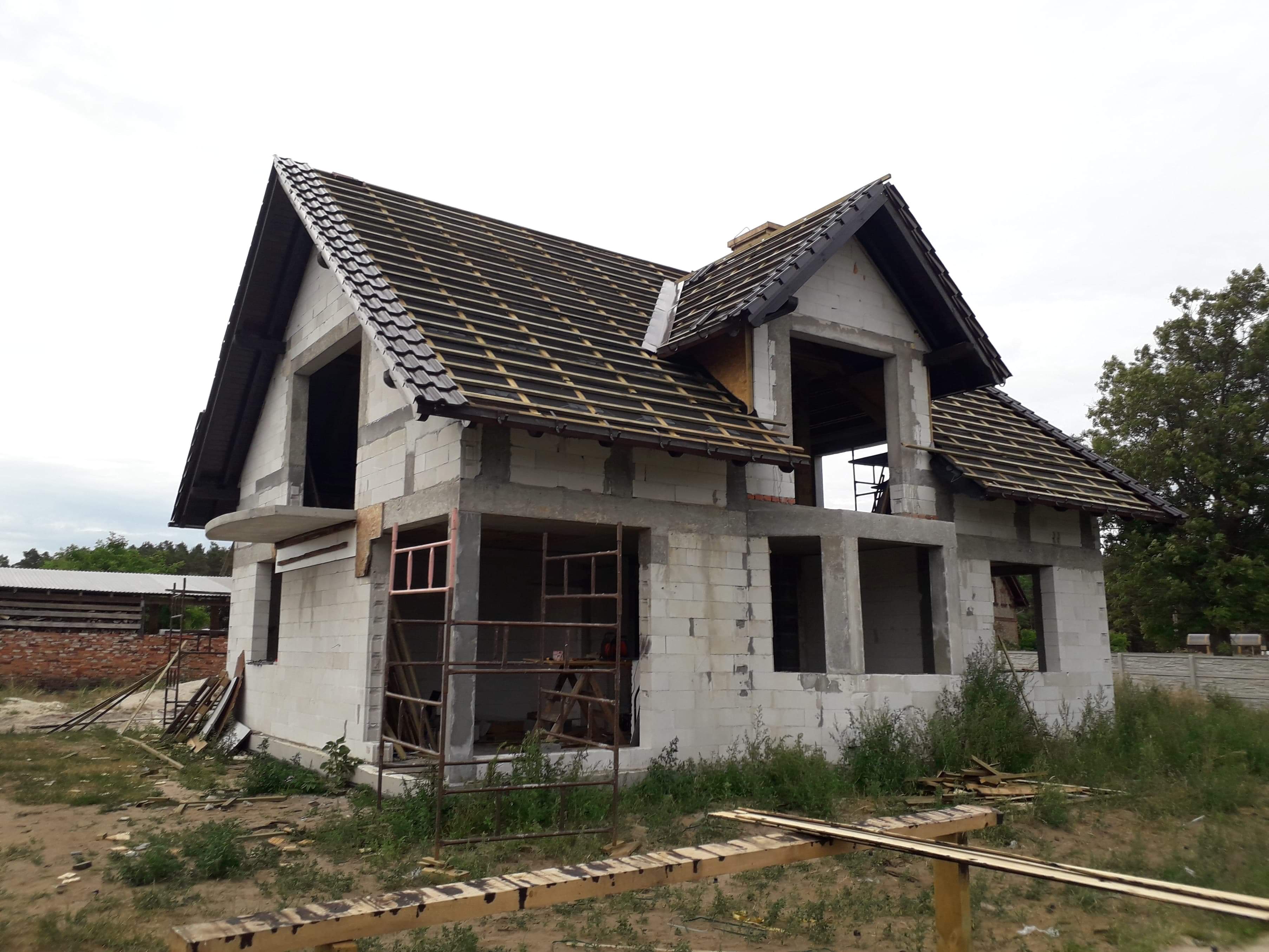 Niedokończony dom z częściowo pokrytym dachem, widoczne krokwie i dachówka, ściany z pustaków, niezadaszony balkon, rusztowania przy oknach, teren zarośnięty trawą.