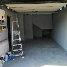 Wnętrze garażu w trakcie remontu, widoczna drabina, puszki z farbą, grzejnik w folii ochronnej, instalacja wodna oraz szare płytki na podłodze.