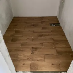 Podłoga w trakcie układania płytek imitujących drewno, z widocznymi krzyżykami dystansowymi, surowymi ścianami i otworem kanalizacyjnym.