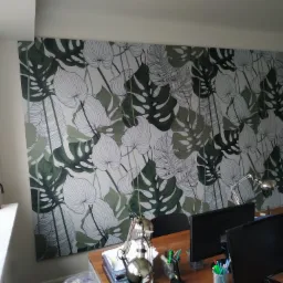 Duża fototapeta z motywem liści monstery i innych roślin tropikalnych na ścianie biura, widok na biurko z dwoma monitorami i lampką, w tle fragment sylwetki mężczyzny.