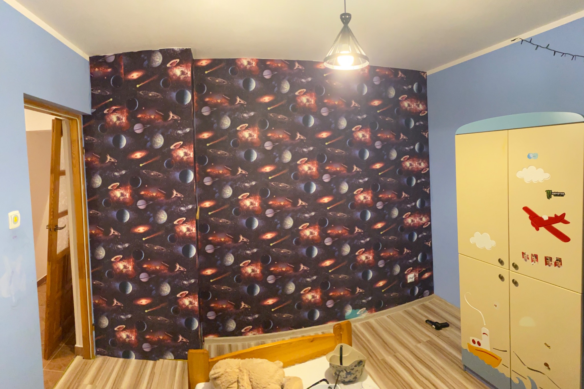 Pokój dziecięcy z tapetą w motywy kosmiczne, drewnianym łóżkiem i szafą z naklejkami samolotów i chmur, oświetlony wiszącą lampą.
