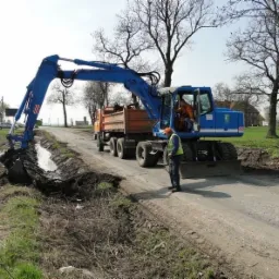 Niebieska koparka na kołach pogłębia rów melioracyjny przy drodze, z pomarańczową ciężarówką w tle i pracownikiem w kamizelce odblaskowej nadzorującym prace.
